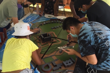 a group of young people huddle around a green architectural model