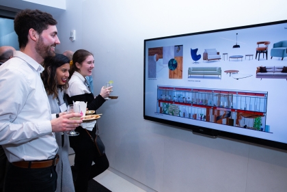Students holding food and beverages smiling and looking at a digital display of architectural designs