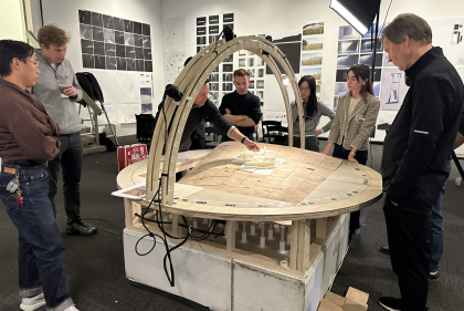 Group of people stand at round, wooden architectural model