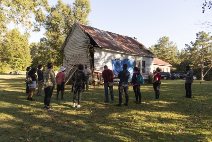 A group of people listen to a man in front of a white wooden structure