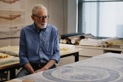 Gray haired bearded man seated at desk with drawing