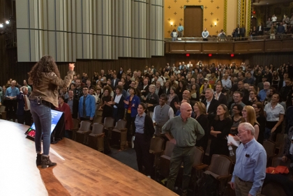 People at auditorium stand and cheer for a young woman speaking on stage