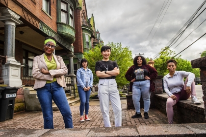 A group of women of various ages and ethnicities pose on the sidewalk outside a victorian home