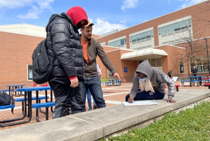 A man gestures to a drawing with two young people outside a brick building