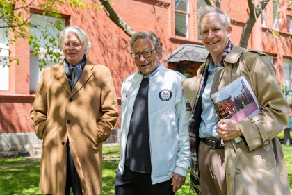 Three men outside a red brick building in dappled sunlight