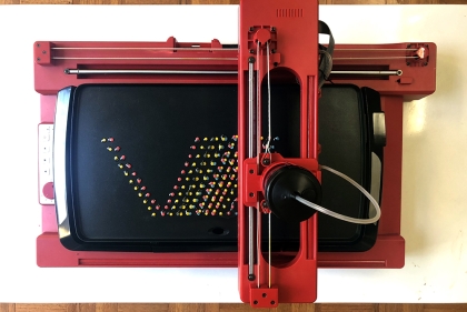 A V-shaped array of rainbow colored dots on black griddle with a red robotic arm above