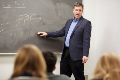 A middle-aged man in a blue blazer in front of a chalk board gesturing 