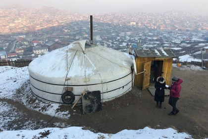 Two women stand outside a white tent dwelling with city in the background
