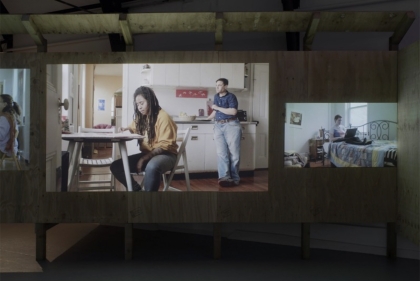 Installation shot of video installation, video showing two people in a kitchen, one of them sitting at a table