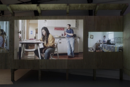 Installation shot of video installation, video showing two people in a kitchen, one of them sitting at a table
