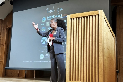 A woman on stage next to a podium gestures at audience 