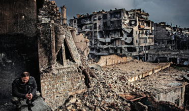 Man squats in foreground of collapsing buildings in Aleppo