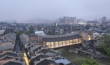 A lantern-like bridge in concrete and wood spans a small river in a dense urban setting