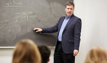 A middle-aged man in a blue blazer in front of a chalk board gesturing 