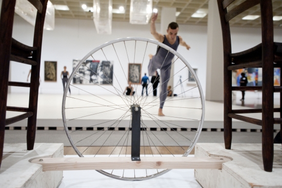 Foreground: sculpture with bicycle wheel suspended between two chairs. Background, performers dancing