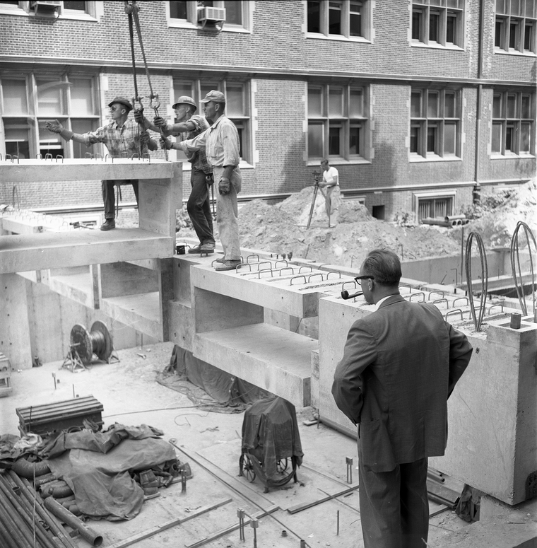 B/W photo of men working at construction site with other man watching them while smoking a pipe