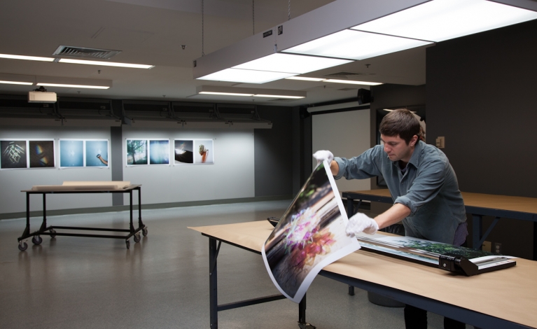 Student prepares to trim a large photograph in a softly lit studio. Large prints of photographs hang on the wall in the back