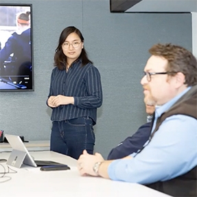 A woman stands in front of a monitor with a man seated in front of her