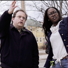 A White man and Black woman outdoors gesture to something outside the frame