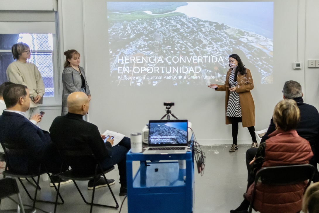 A young woman in front of a projector screen addresses a seated audience