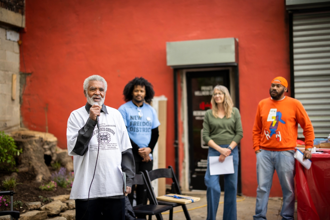 An older man in a t-shirt speaks into a mic with three people behind listening