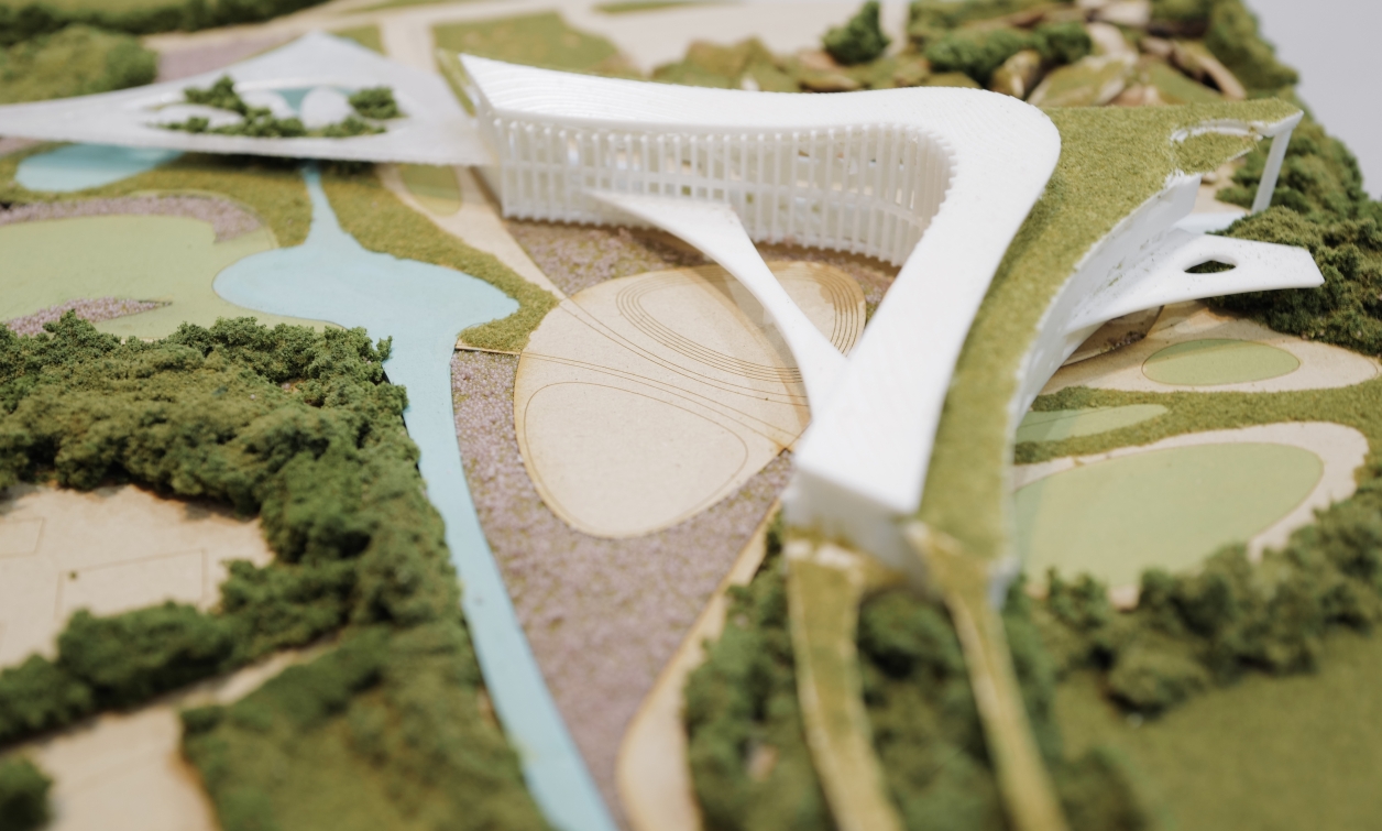 Photo of a physical site model for a school project, highlighting the Y-shaped architecture integrated with curved landscape paths, water features, and surrounding forested areas. Emphasis on form merging with terrain.
